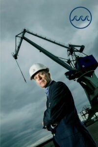 Kim Wikström står framför en lyftkran. Bakgrunden är hotfullt grå. I det övre högra hörnet syns en logo bestående av tre vågor inne i en cirkel.